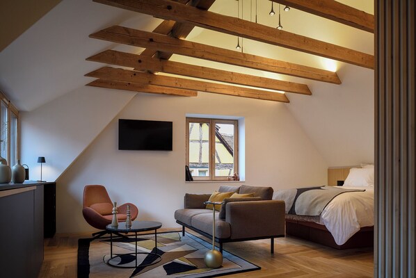 A modern loft bedroom with exposed wooden beams, a large window, a bed, a sofa, and a desk with a chair.