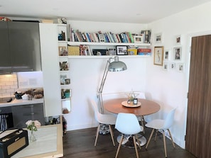 Une cuisine compacte avec un coin repas, comprenant une table ronde entourée de quatre chaises, une suspension et une bibliothèque remplie de livres et d’objets décoratifs.