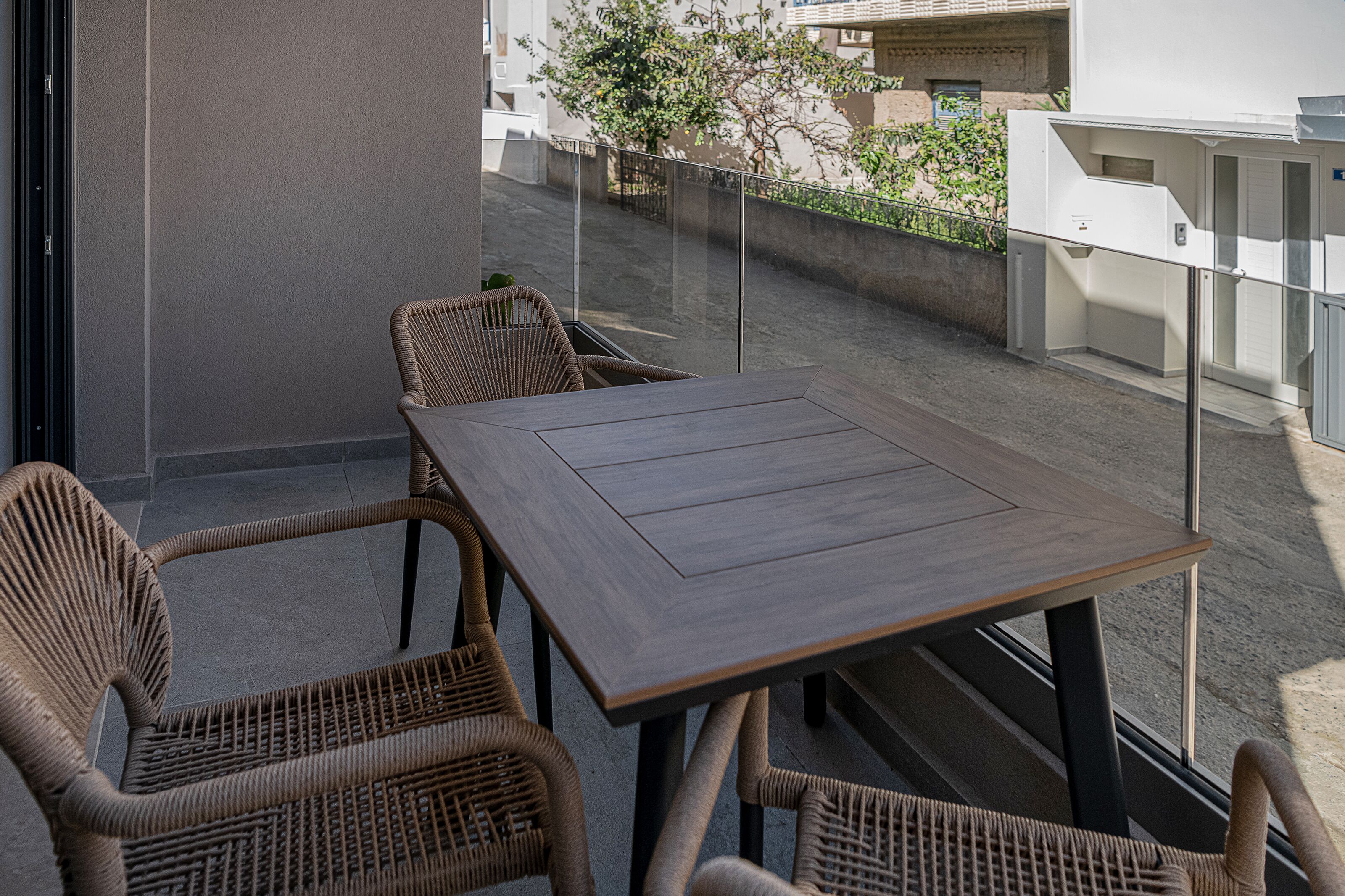 A balcony with wicker furniture and a wooden table, overlooking a residential area with a low wall and greenery.