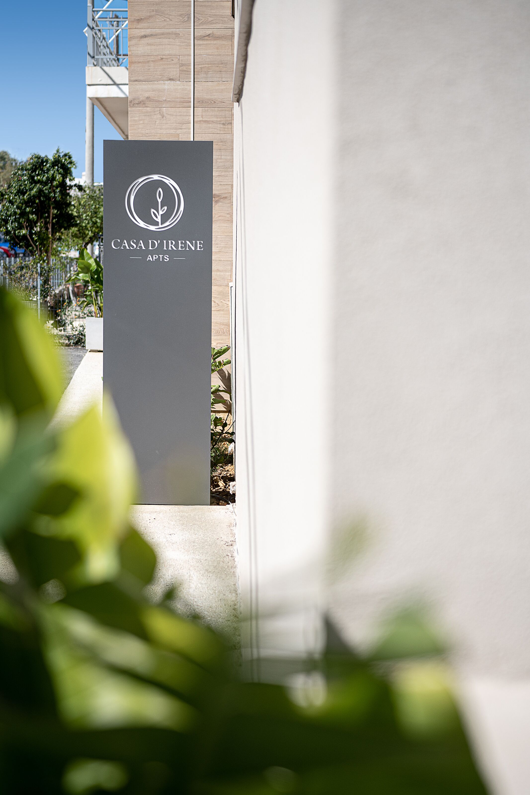 A sign for Casa d'Irene Apts with a modern design, located at the entrance of a building with a concrete pathway and greenery.