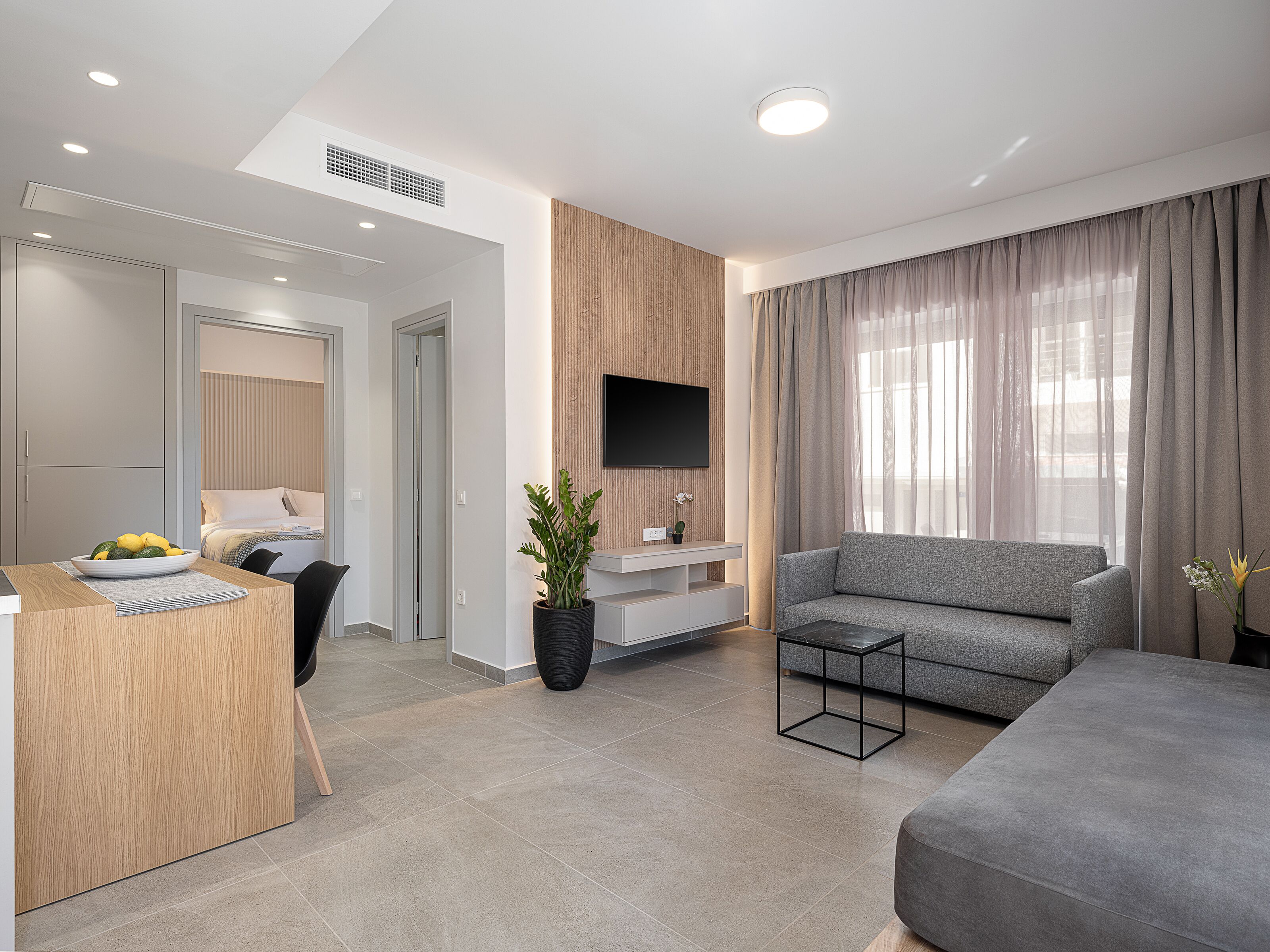Una sala de estar moderna con un sofá gris, un comedor con una mesa de madera, y un televisor montado en la pared.