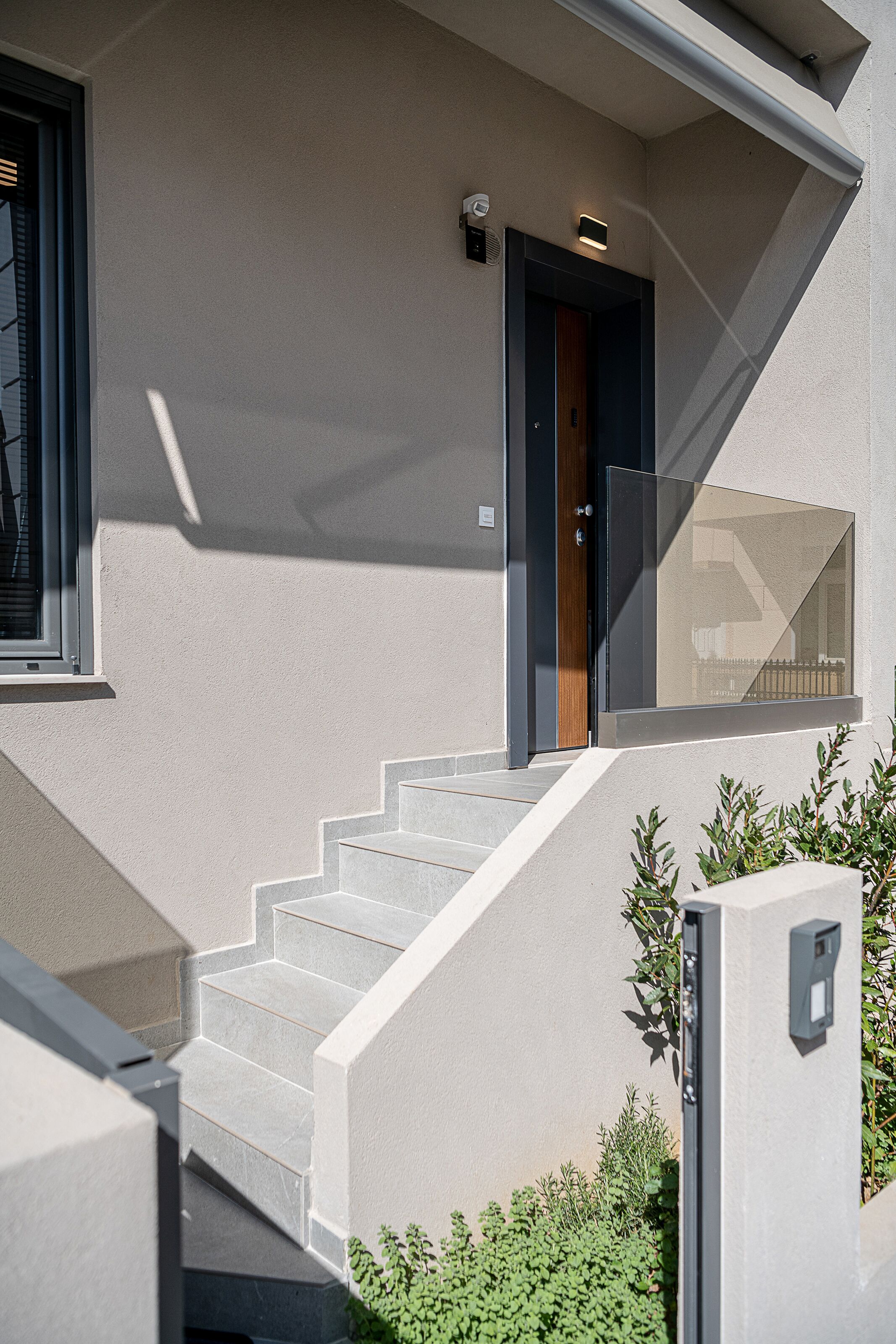 Una casa moderna con una escalera que lleva a una puerta oscura, una ventana con persianas y un pequeño espacio exterior con plantas.