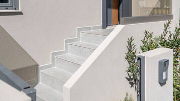 Una casa moderna con una escalera que lleva a una puerta oscura, una ventana con persianas y un pequeño espacio exterior con plantas.