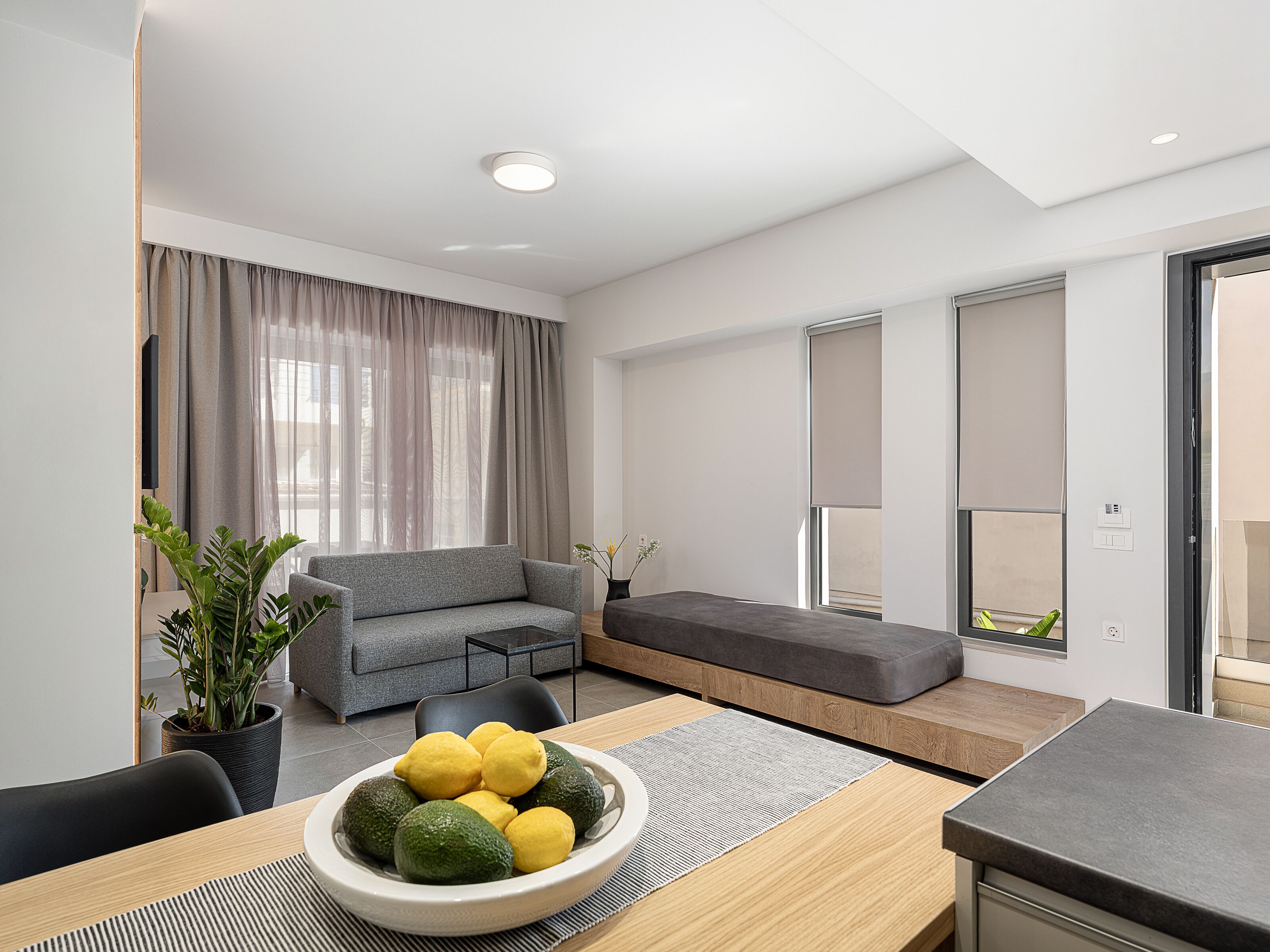 Una sala de estar moderna con un sofá gris, una mesa de comedor con un cuenco de fruta y un ventanal con cortinas.