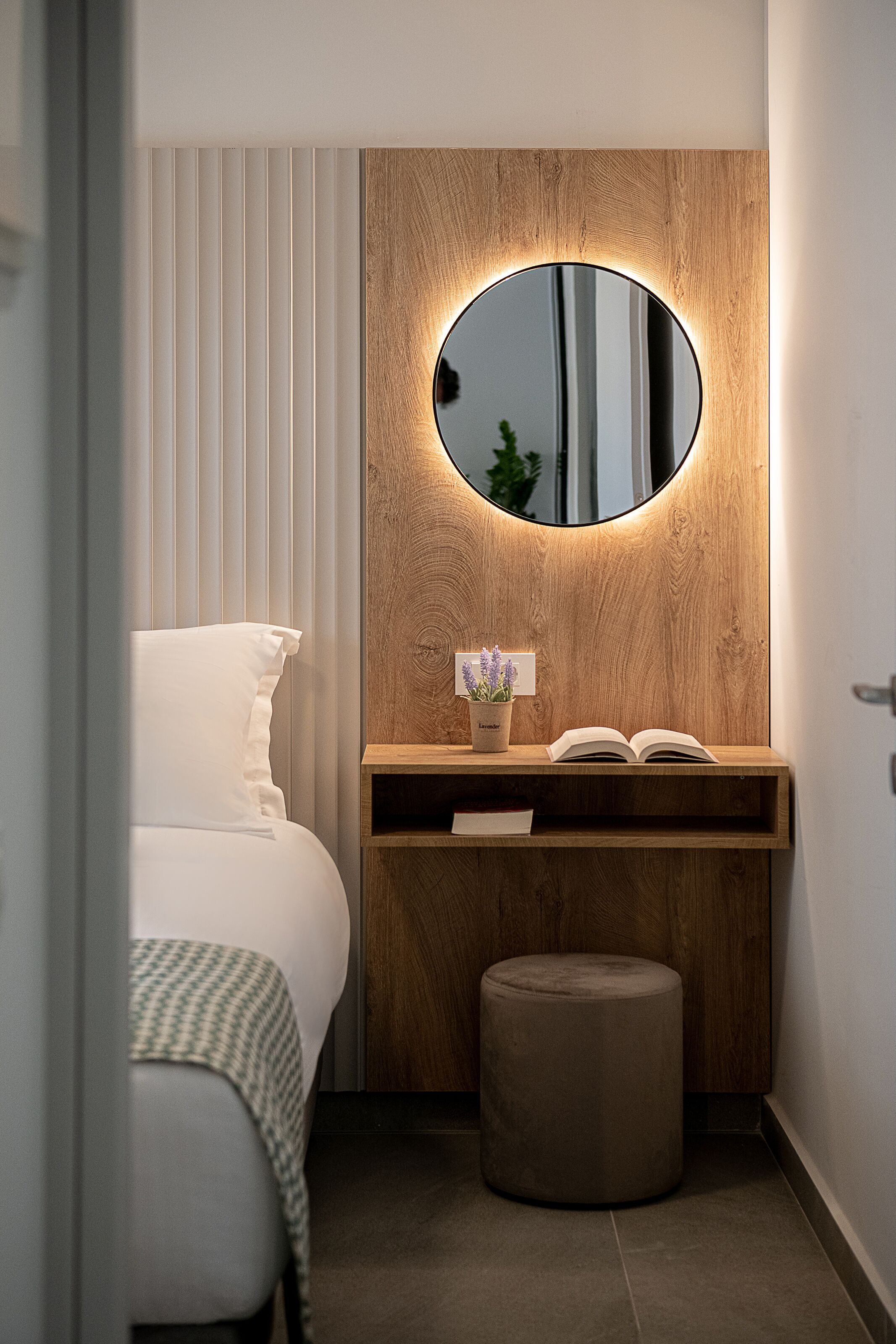 A modern bathroom with a wooden vanity, a round mirror, a small potted plant, and a bedside table with an open book.