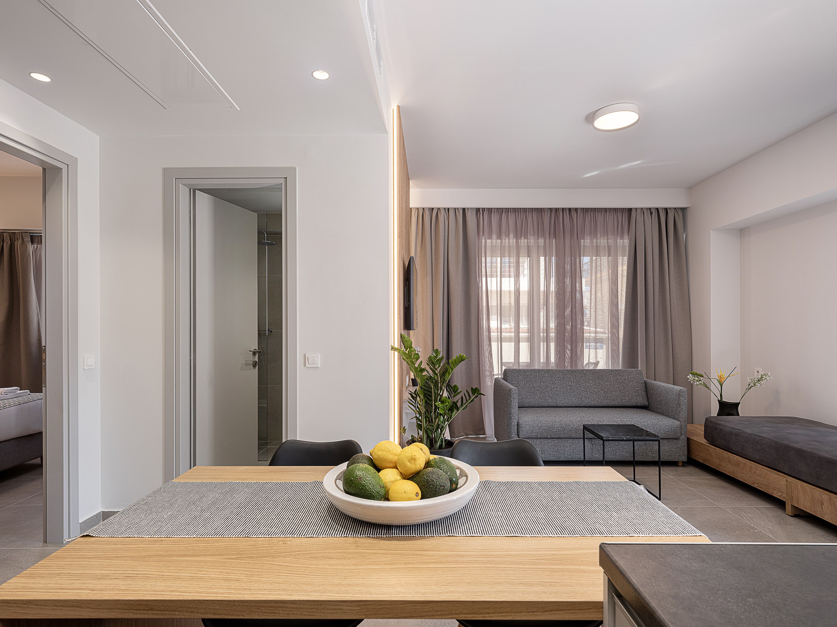 A modern living room with a grey sofa, a dining table with a bowl of fruit, and a view of the outside through large windows.