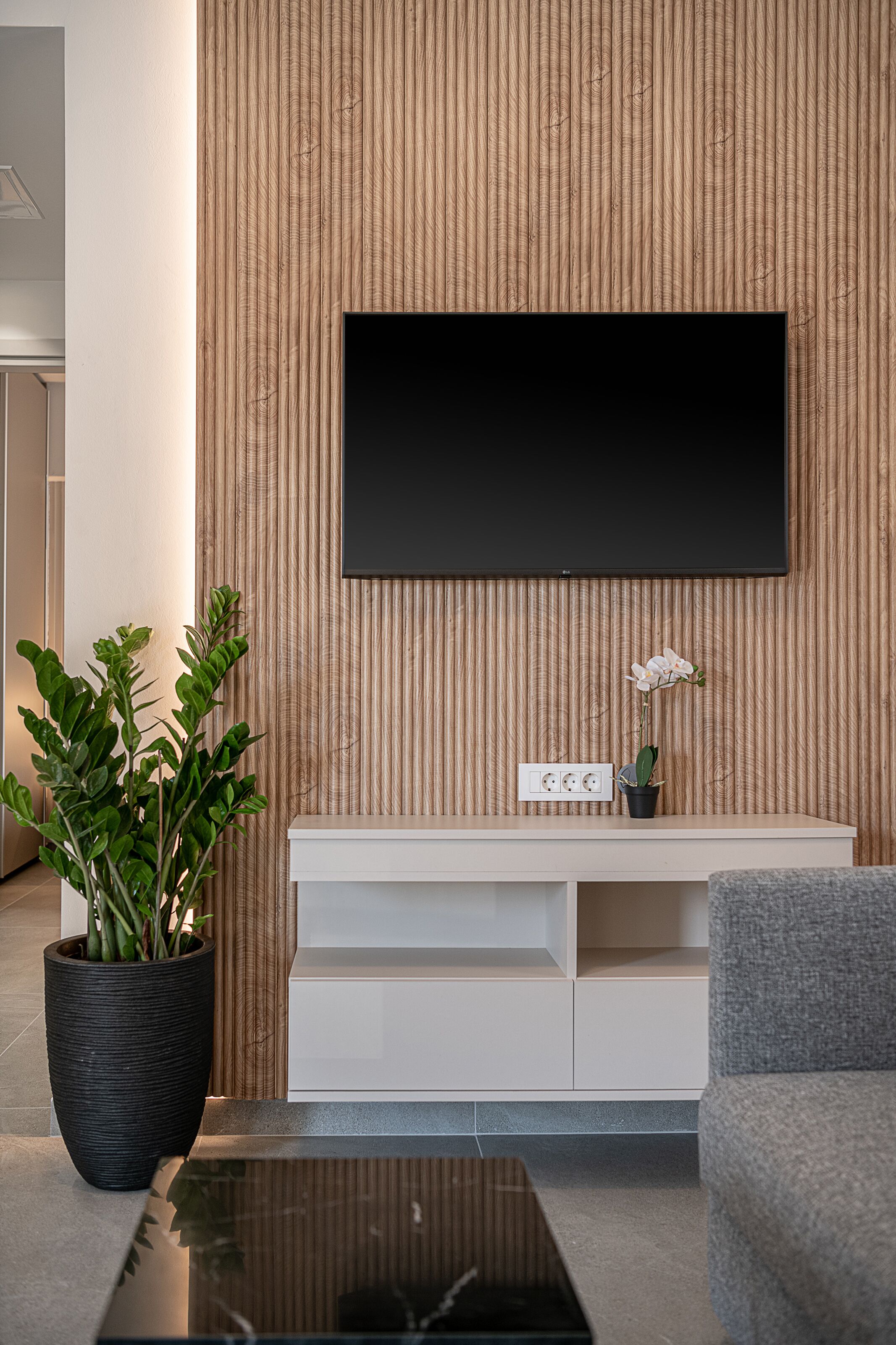 Een moderne woonkamer met een flatscreen televisie aan een houten wand, een wit tv-meubel, een grijze bank en een grote potplant.