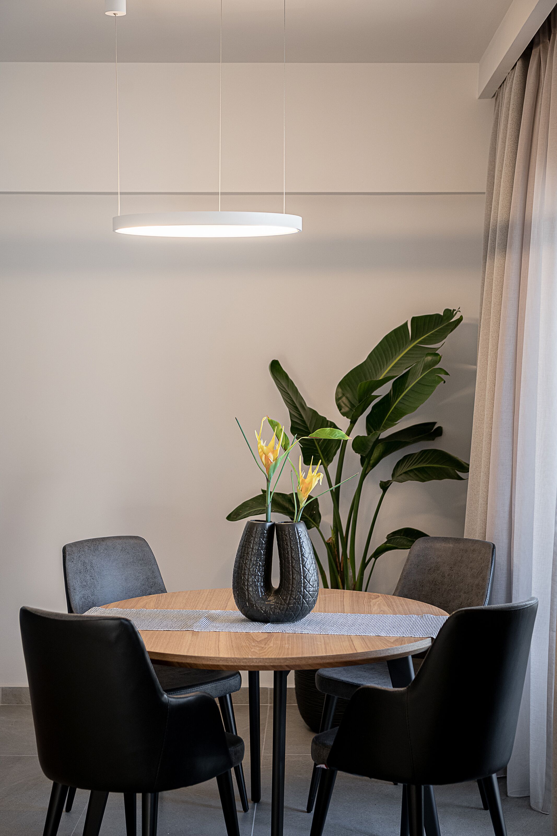 Ein moderner Essbereich mit einem Holztisch, schwarzen Stühlen, einer Vase mit gelben Blumen und großen Grünpflanzen.