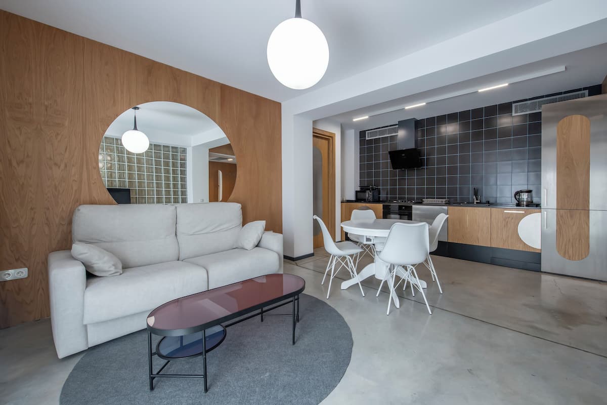 A modern living room with a sofa, coffee table, and dining area with chairs. There is a kitchen in the background with a TV and a refrigerator.