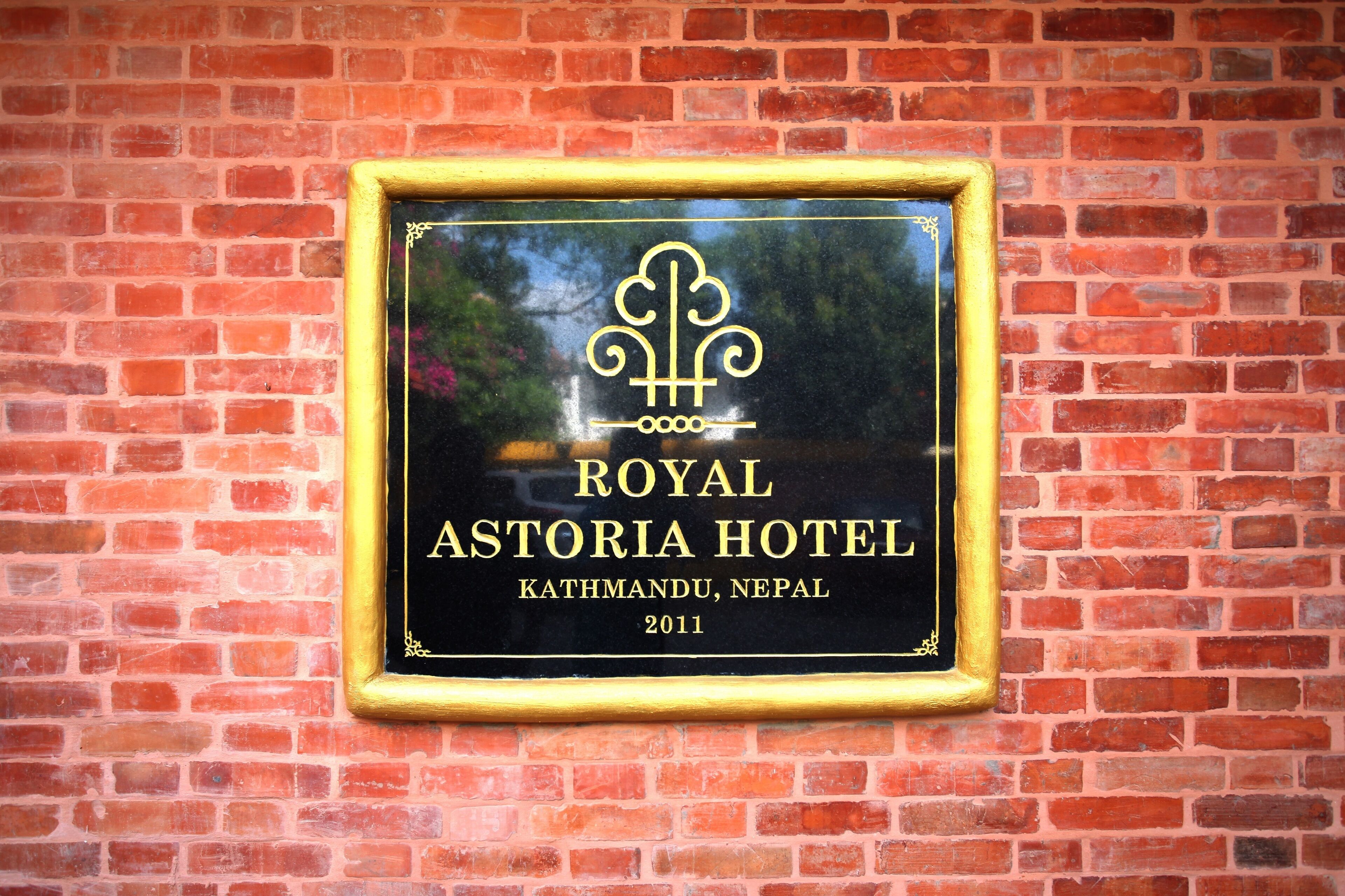 Une plaque en métal fixée à un mur de briques, portant l’inscription : « ROYAL ASTORIA HOTEL, KATHMANDU, NÉPAL 2011 ».