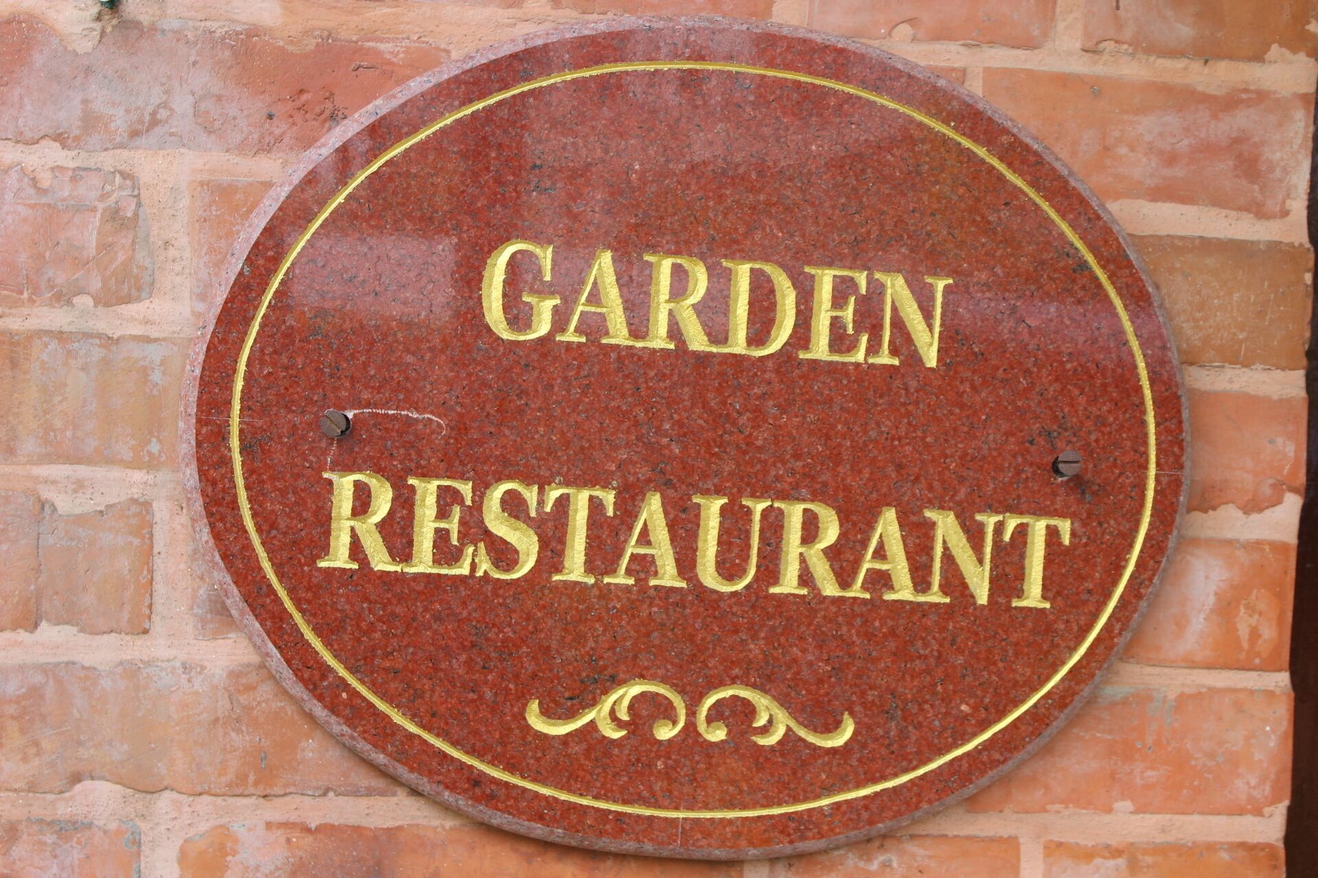 Een rond bord met de woorden 'GARDEN RESTAURANT' in gouden letters op een rode achtergrond, bevestigd aan een bakstenen muur.
