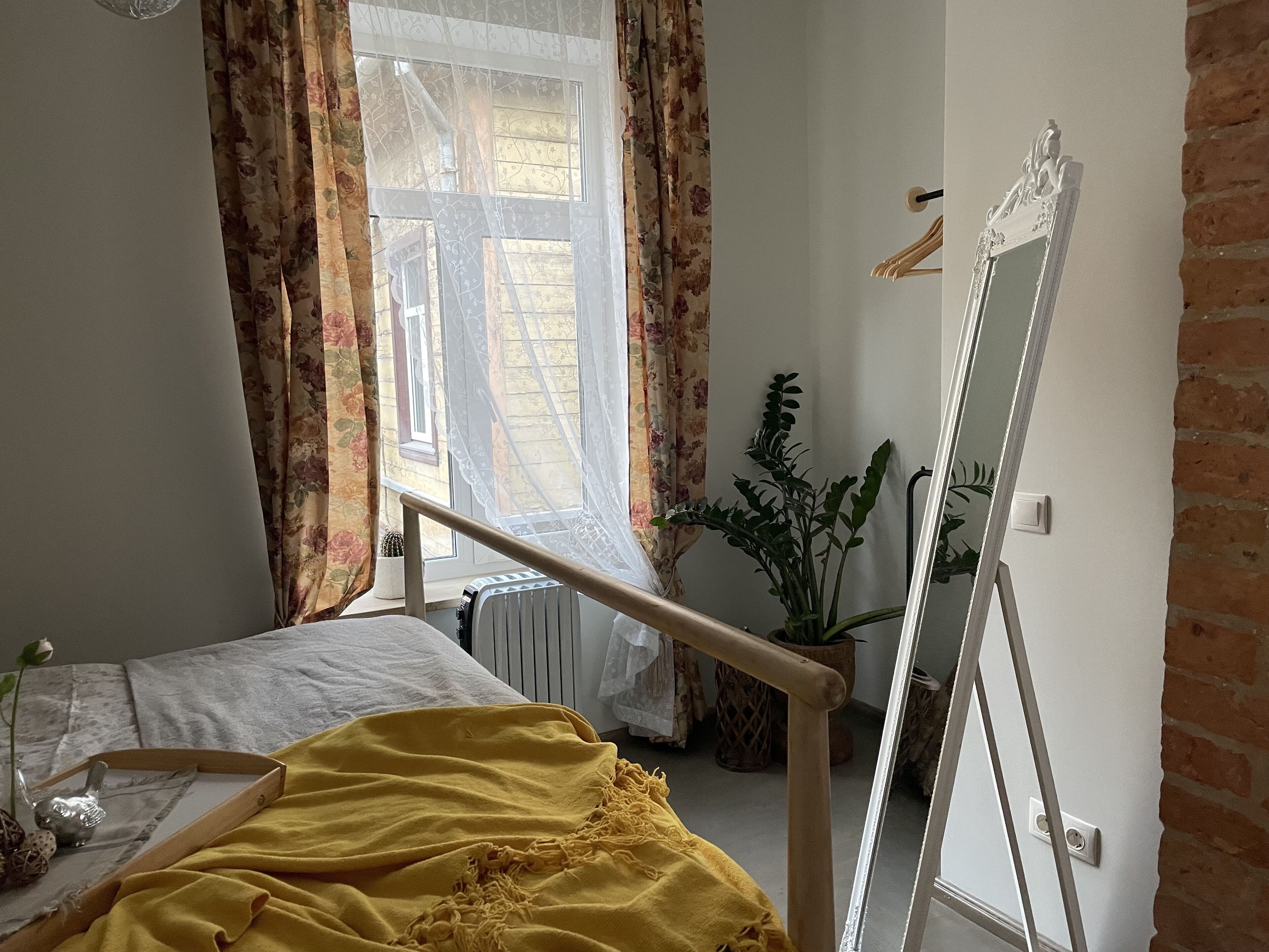 Une chambre à coucher avec un lit, une fenêtre avec des rideaux, un radiateur, une plante en pot et un miroir.