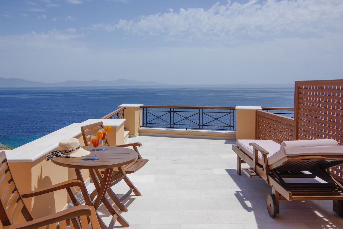 A sunlit outdoor seating area with a view of the sea, a table with drinks, and lounge chairs.