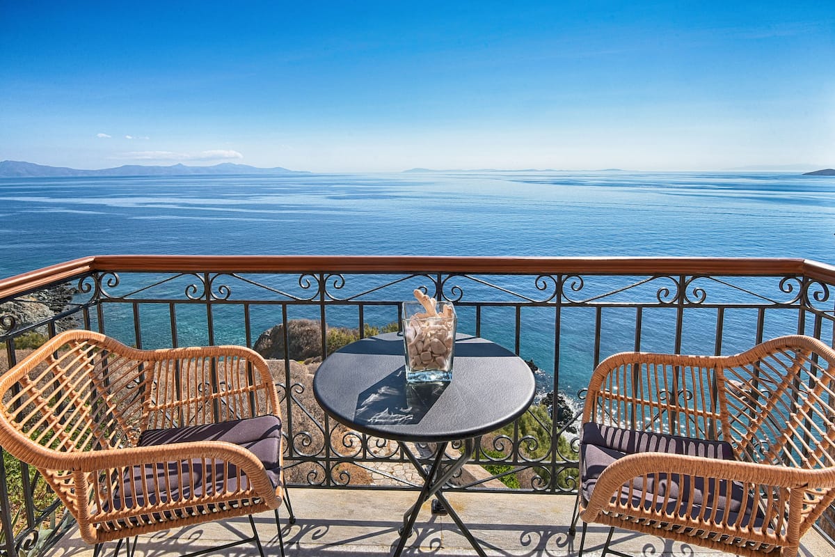 A balcony with wicker chairs and a small table, overlooking a vast body of water and distant mountains.