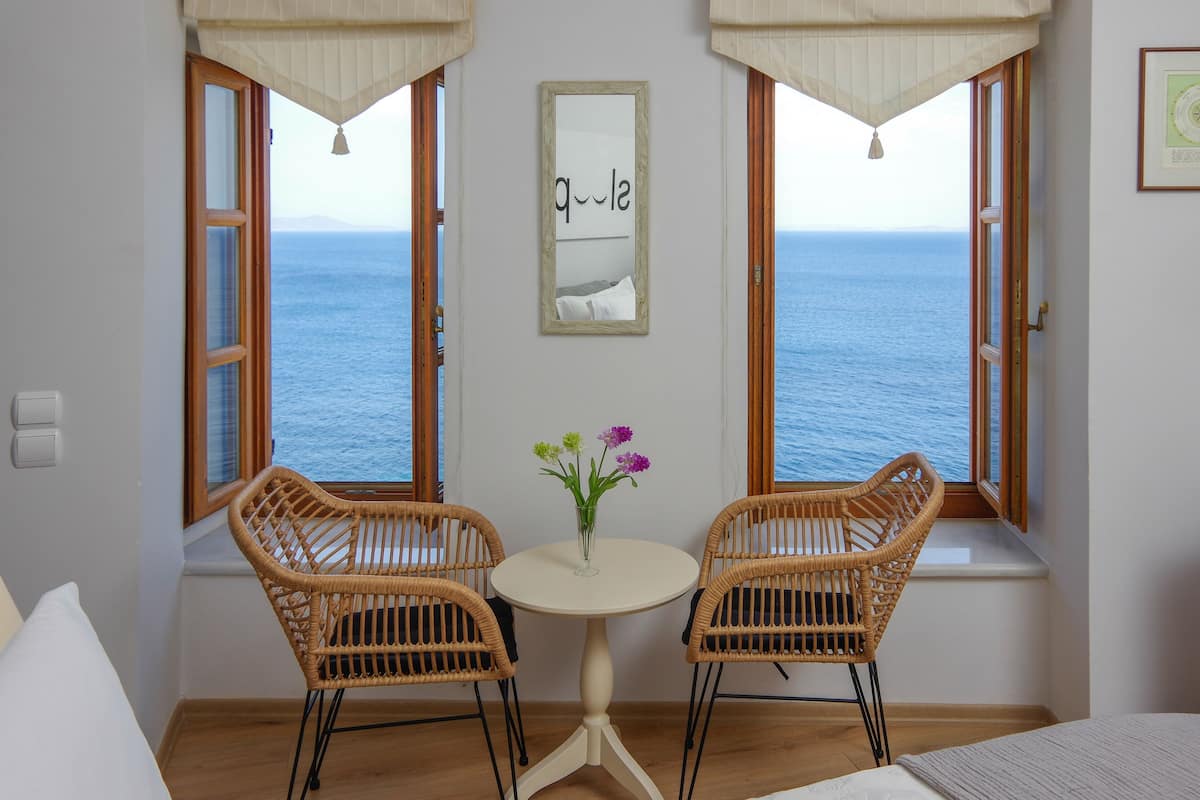 A room with a view of the sea, two wicker chairs, a small table with a vase of flowers, and a framed picture on the wall.