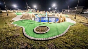 Een goed verlichte minigolfbaan 's nachts, met een fontein in het midden en beplante omgeving.