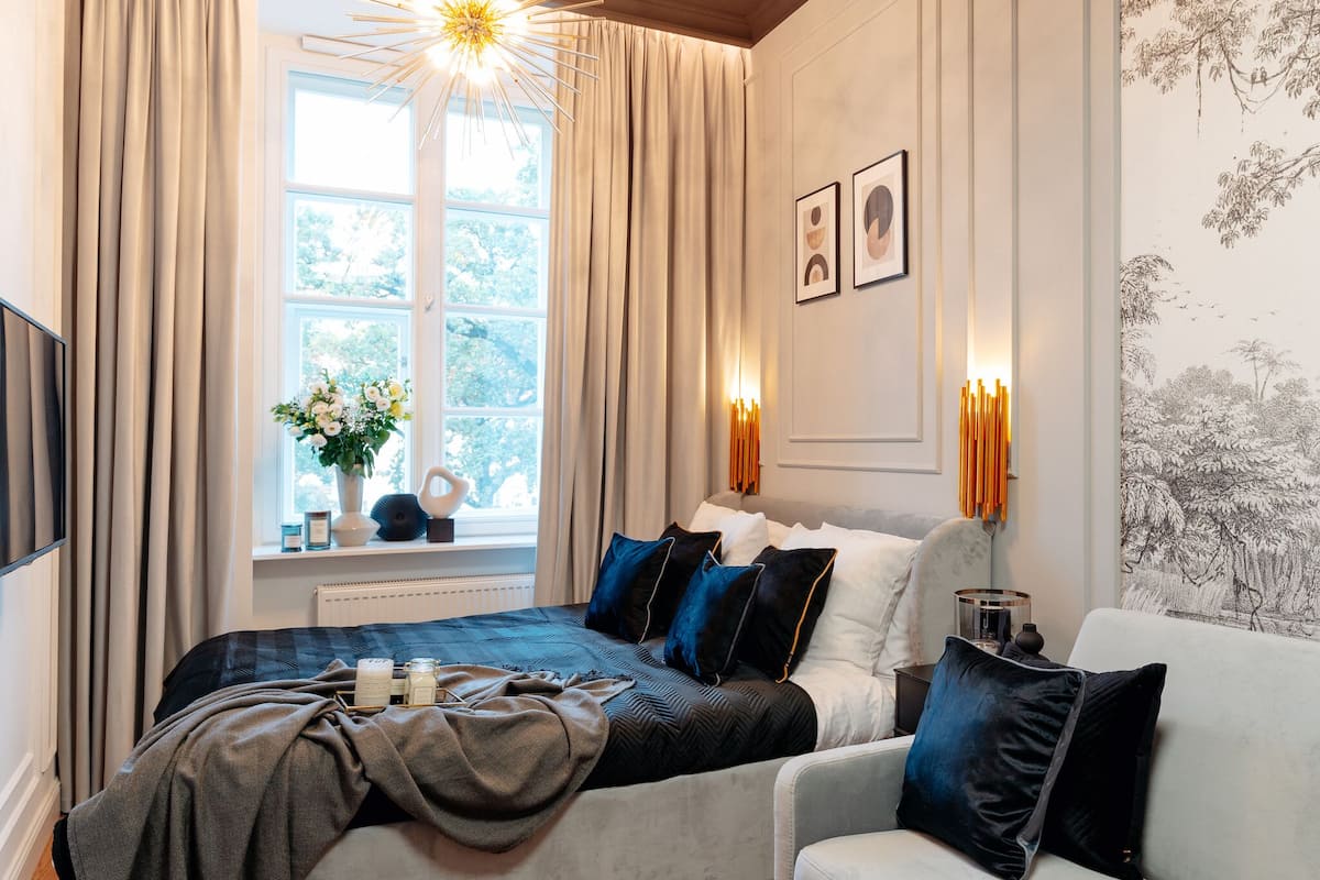 Un dormitorio con cama, ventana con cortinas, mesita de noche con un jarrón y velas, y una pared decorada con cuadros.