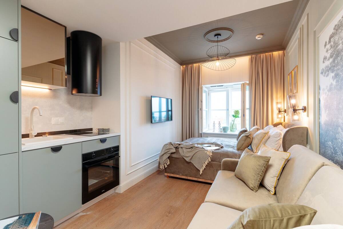 Una sala moderna con un sofá, un televisor de pantalla plana, una zona de cocina con estufa y un ventanal con cortinas.