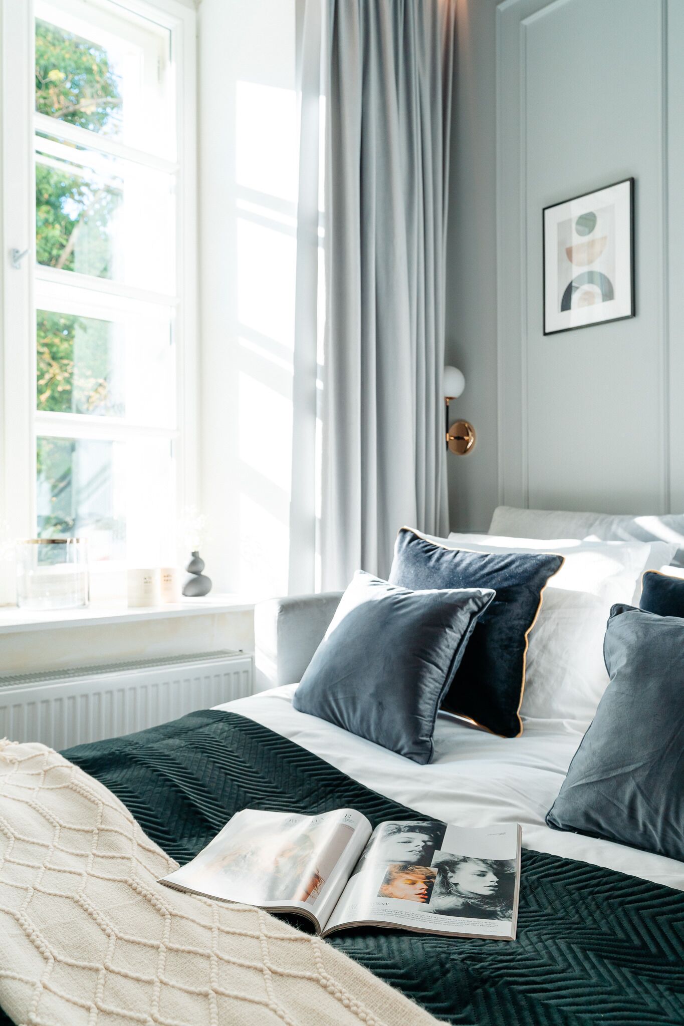 Una cama bien hecha con almohadas oscuras y claras, una revista sobre la cama y una ventana con cortinas.