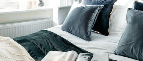 Una cama bien hecha con almohadas oscuras y claras, una revista sobre la cama y una ventana con cortinas.