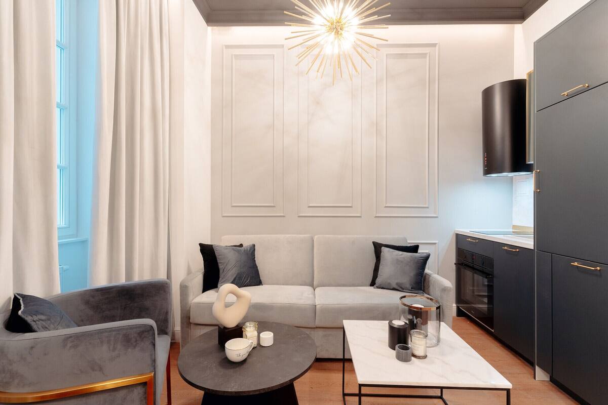 Una sala de estar moderna con un sofá gris, una mesa redonda de centro y una cocina con armarios oscuros y horno empotrado.
