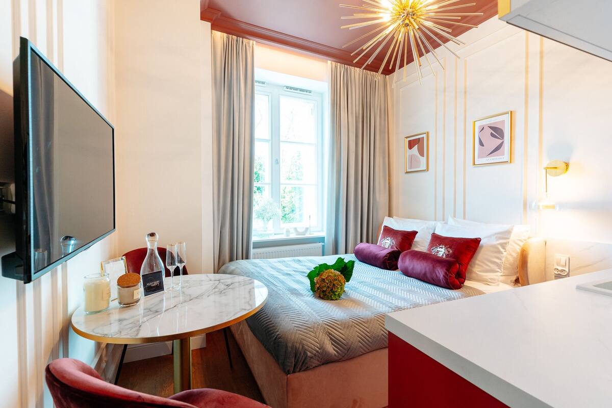 Una habitación de hotel con cama, televisión, una mesa con vela y vasos, y ventana con cortinas.