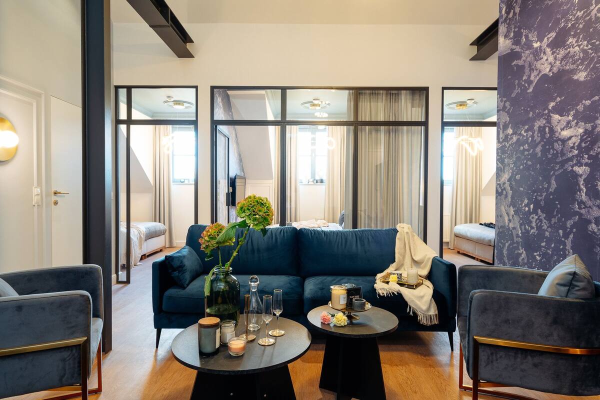Una sala moderna con un sofá azul oscuro, dos sillones, una mesita de centro con velas, un jarrón con flores y un espejo grande que refleja la habitación.
