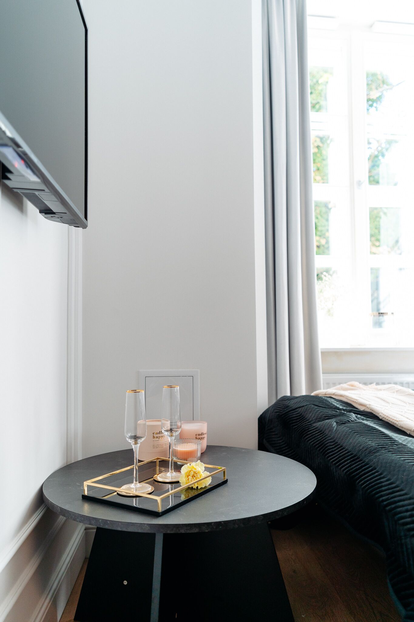 Eine Ecke eines Zimmers mit einem runden Tisch, auf dem ein Tablett mit Kerzen und Gläsern steht, einem dunklen Sofa und einem Fenster mit Vorhängen.