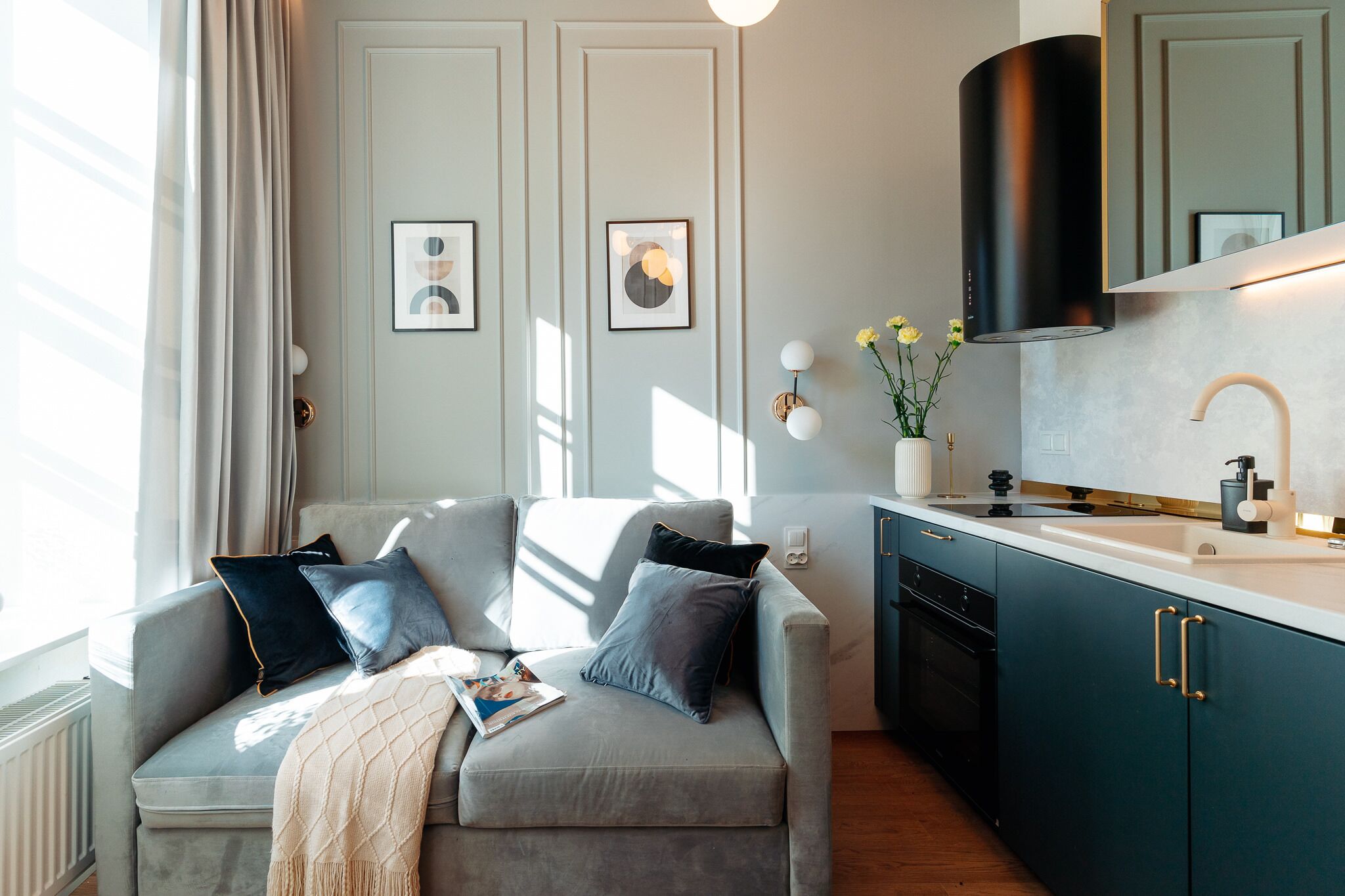 A modern living room with a grey sofa, a small table with a magazine, and a kitchen area with teal cabinets and a black range hood.