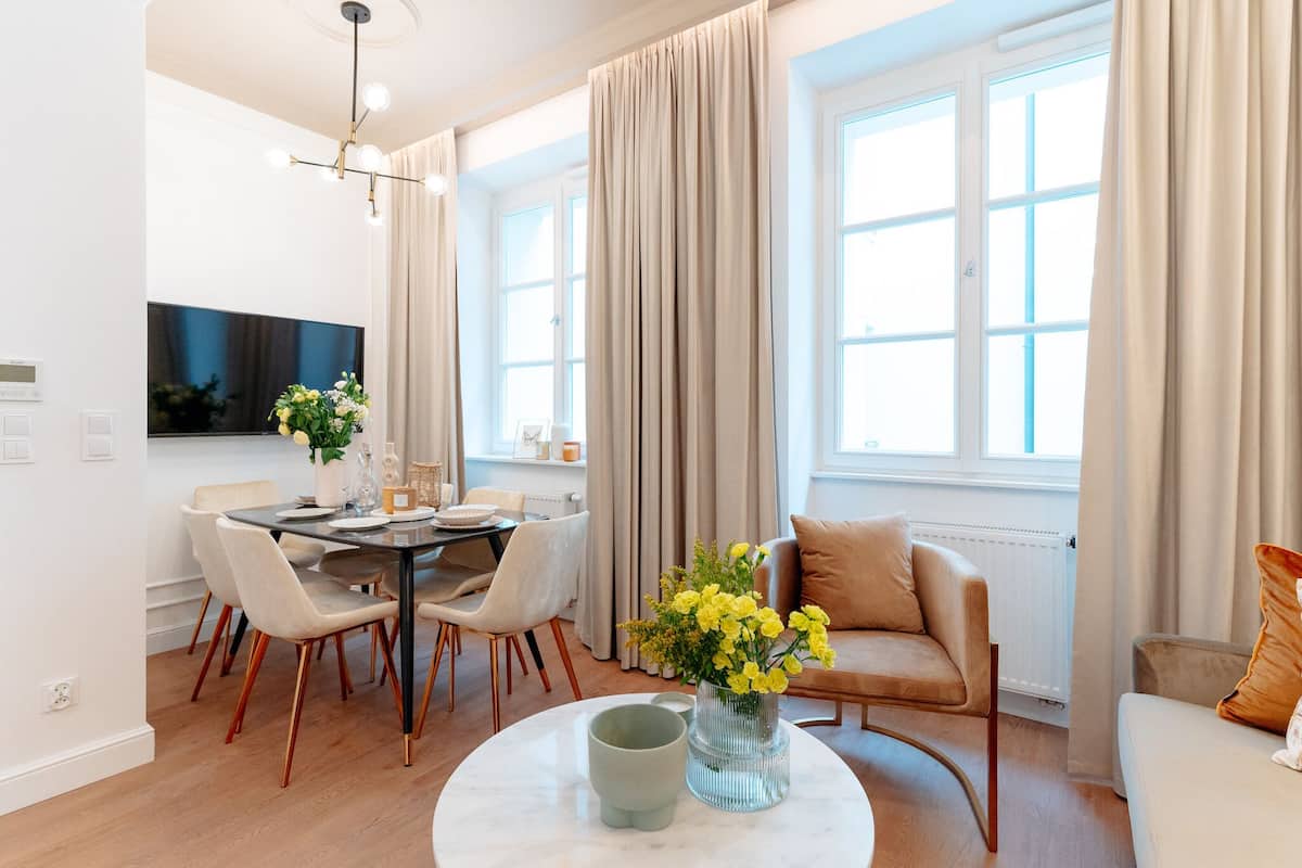 Una sala de estar moderna con comedor, ventanas grandes con cortinas, un televisor de pantalla plana y un florero con flores sobre una mesa de centro.