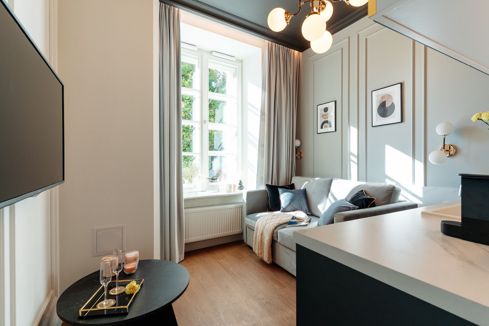 A modern living room with a sofa, a coffee table, and a TV. There is a window with curtains, a radiator, and a wall-mounted light fixture.