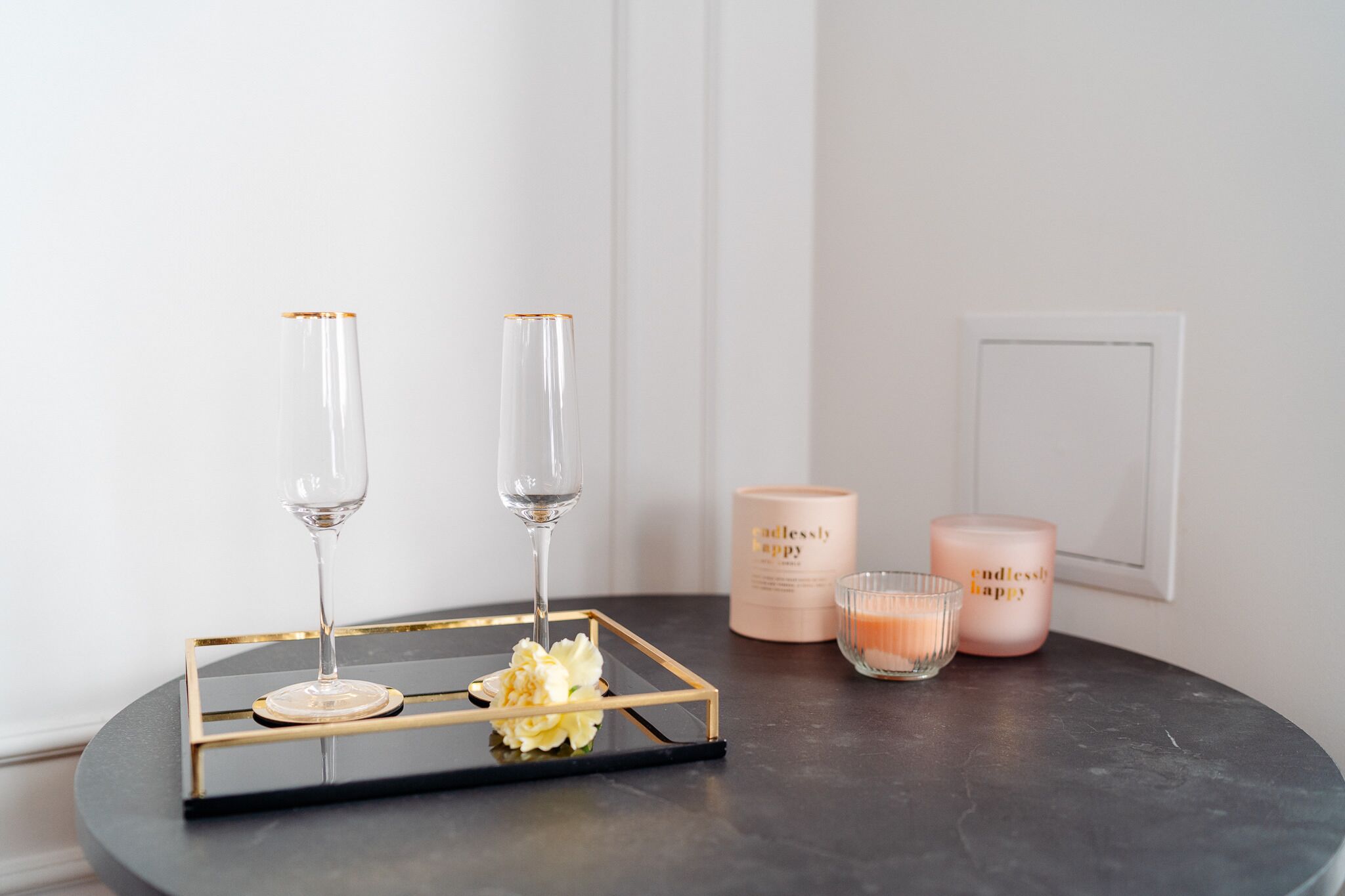 A table with two glasses of water, a tray with a flower, and two candles on a dark surface.
