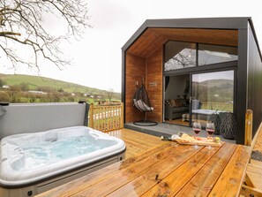 A wooden deck with a hot tub, a dining table with food and drinks, and a view of the countryside. - Doe Lodge (Llandrindod Wells)