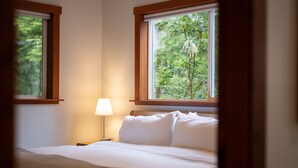 Een net opgemaakt bed met witte lakens en een beige plaid, geplaatst onder een raam met uitzicht op groen.