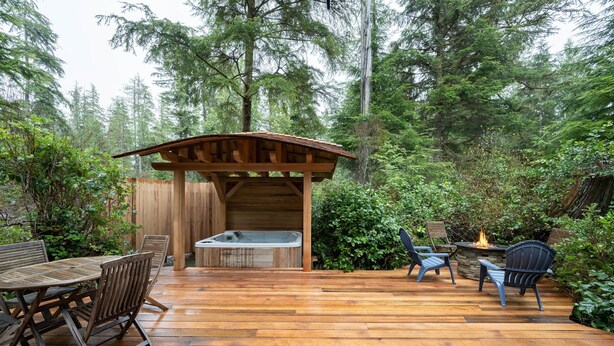 Een houten terras met een jacuzzi, buitenmeubels en een vuurkuil, omringd door dicht bos.