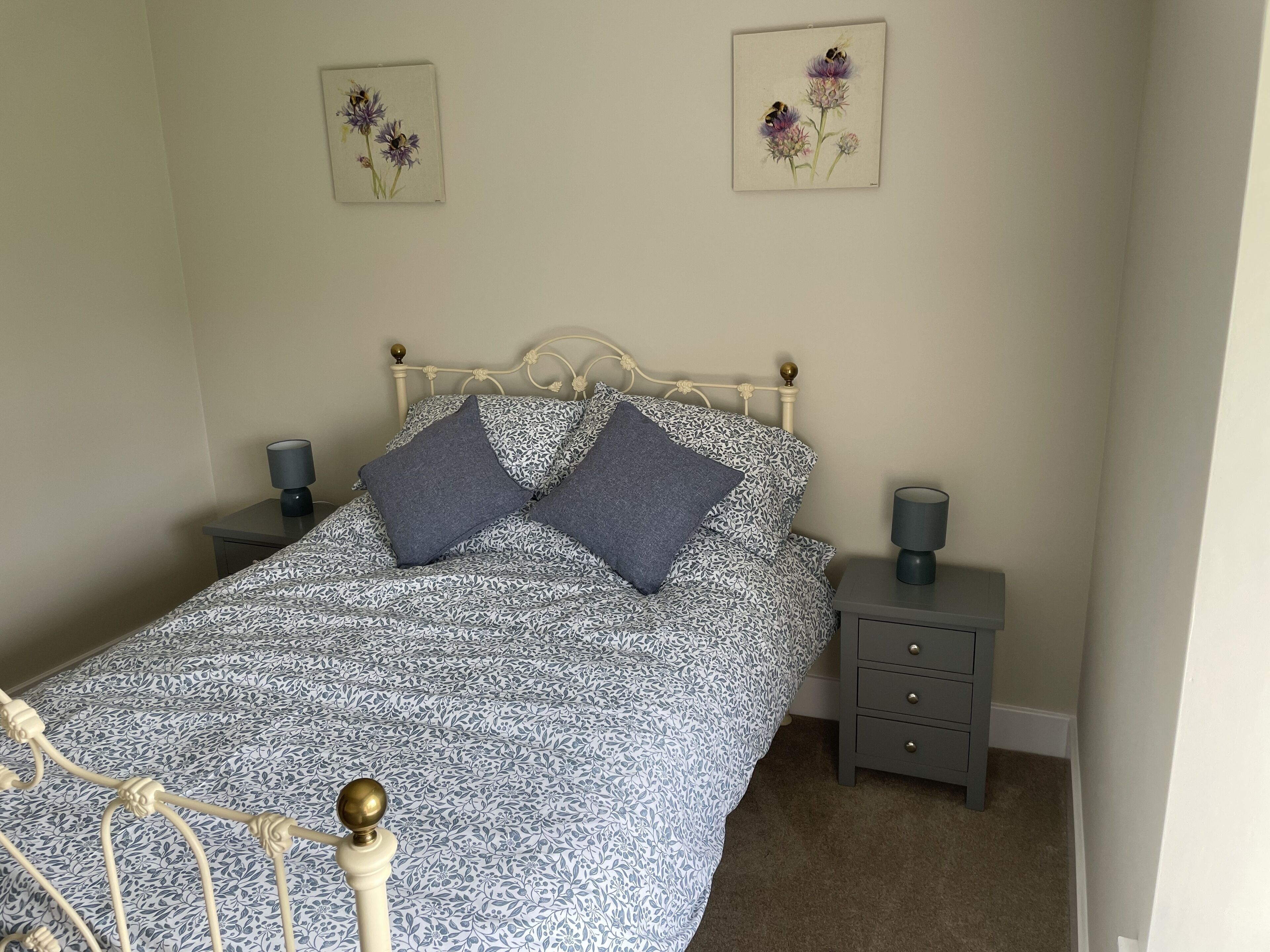 A neatly made bed with a patterned bedspread, two pillows, a metal headboard, a bedside table with a lamp, and a small drawer.