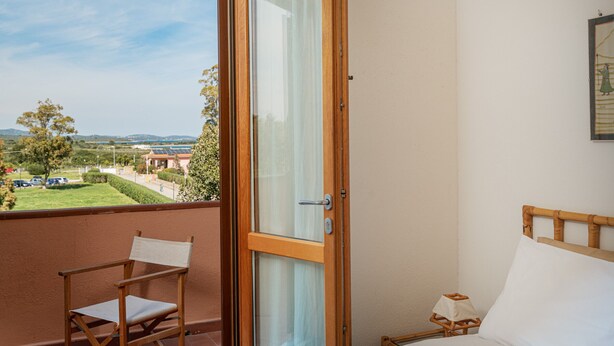 Un balcon avec une chaise en bois, un lit avec une tête de lit en bois et une vue sur un paysage verdoyant avec des bâtiments visibles au loin.
