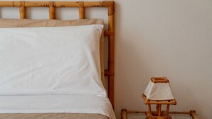 Un lit avec une tête de lit en bambou, un oreiller blanc et une petite table accompagnée d’une chaise en bambou.