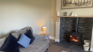 A living room with a grey sofa, a lit table lamp, a fireplace with a stone surround, and a painting above the mantel. - Till Cottage 3 Fenton Hill Farm (Wooler)