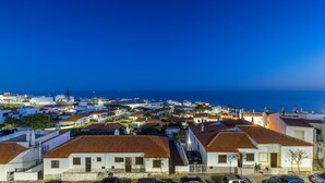 Un pueblo costero al anochecer con edificios blancos y tejados de tejas rojas, una vista despejada del mar, y coches estacionados en primer plano.
