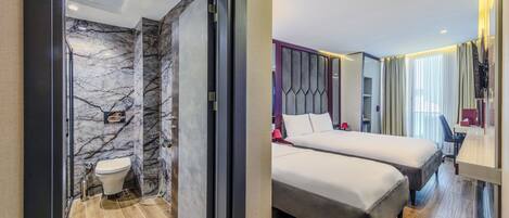 Una habitación de hotel moderna con un baño de mármol, dos camas, un escritorio y un ventanal con cortinas.