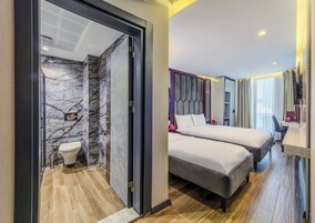 Una habitación de hotel moderna con un baño de mármol, dos camas, un escritorio y un ventanal con cortinas.