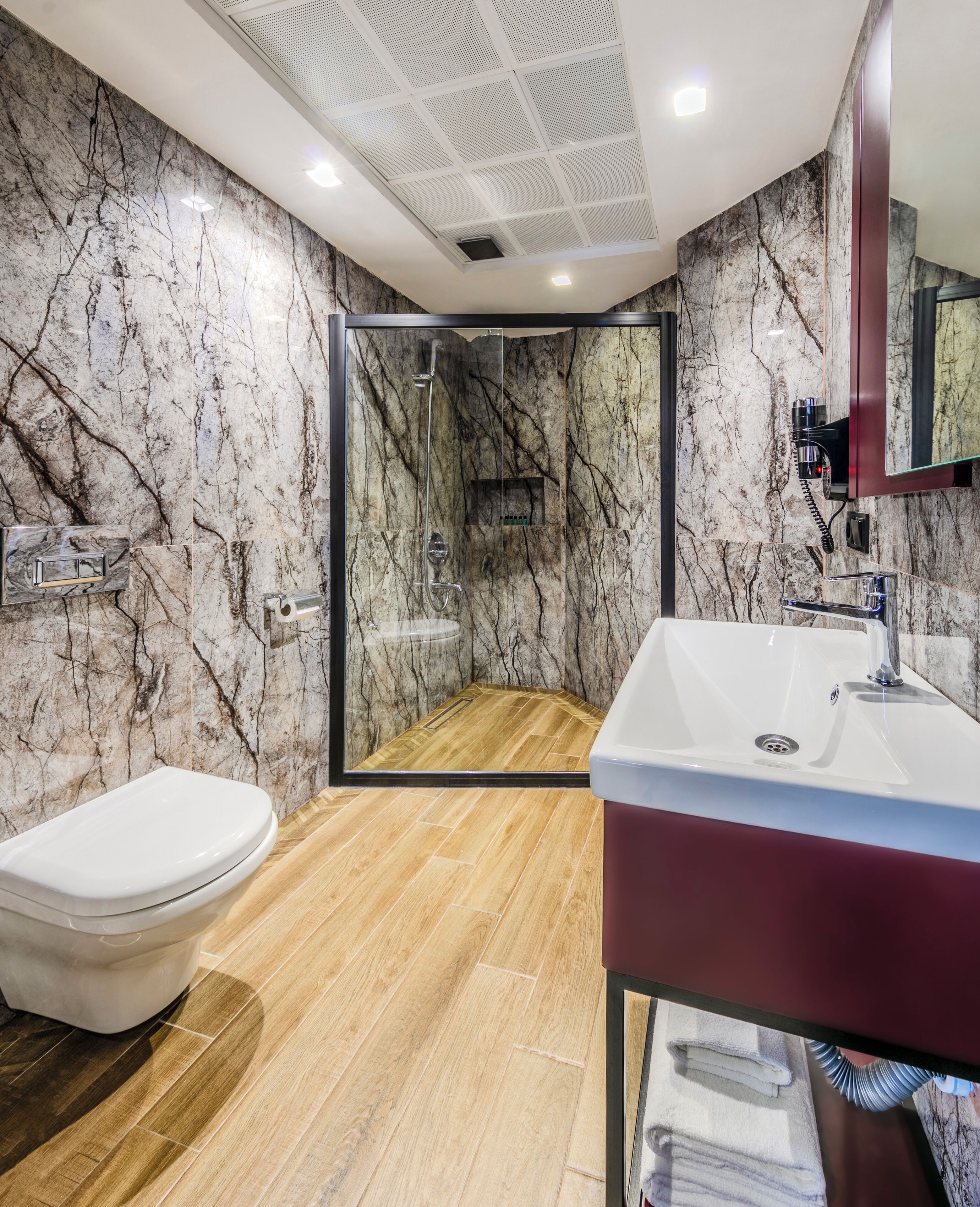 A modern bathroom with a marble wall, a wooden floor, a white toilet, a sink with a red basin, and a glass shower enclosure.