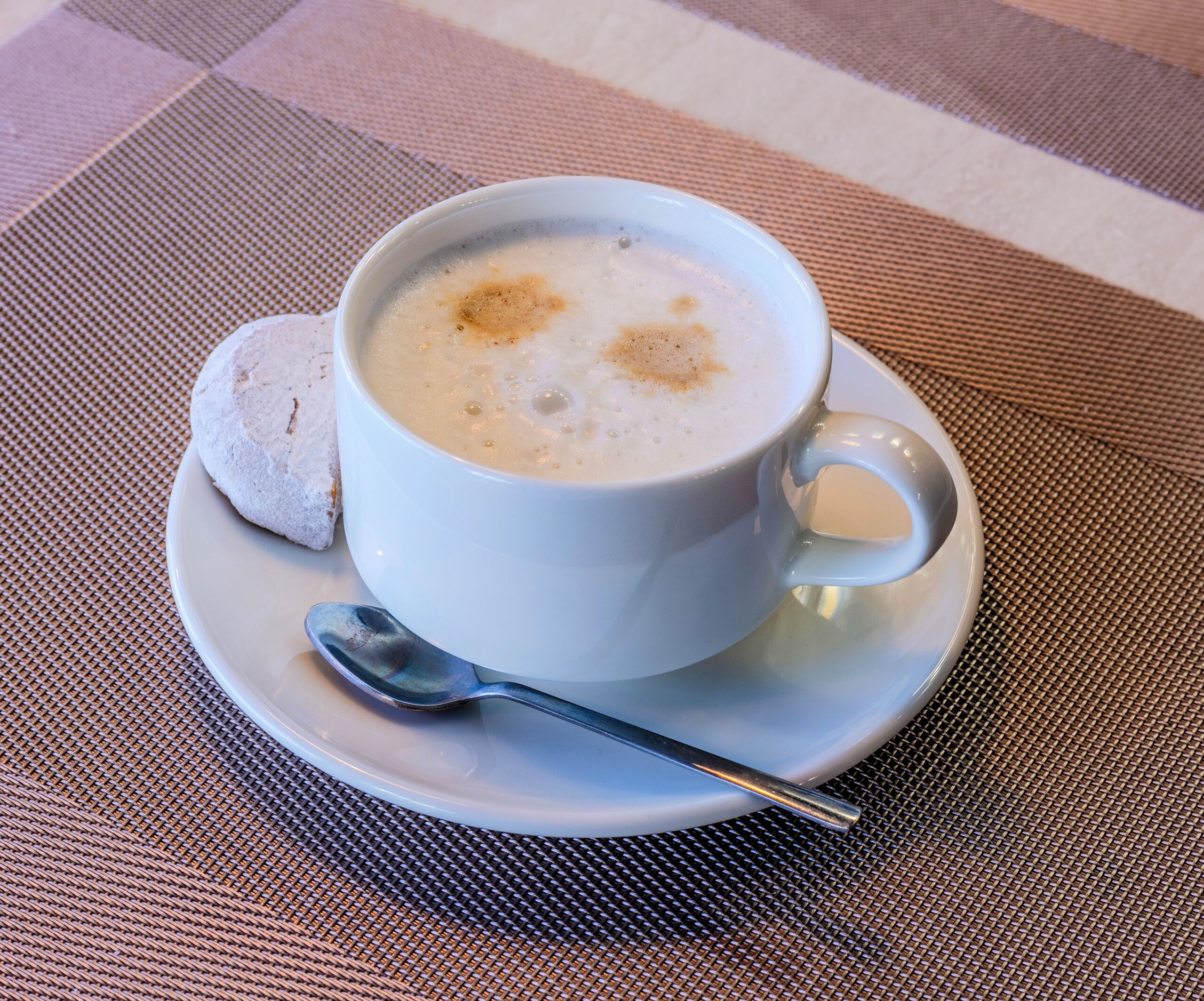 Une tasse de café blanc, surmontée d’une mousse, posée sur une soucoupe blanche, avec une cuillère à côté, le tout sur un set de table en fibres tressées.