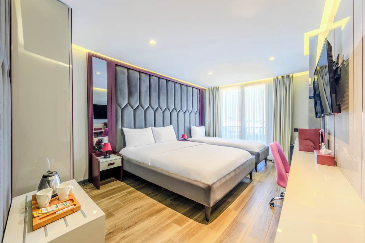 A modern hotel room with a large bed, a bedside table with a lamp, a TV, and a desk with a chair.