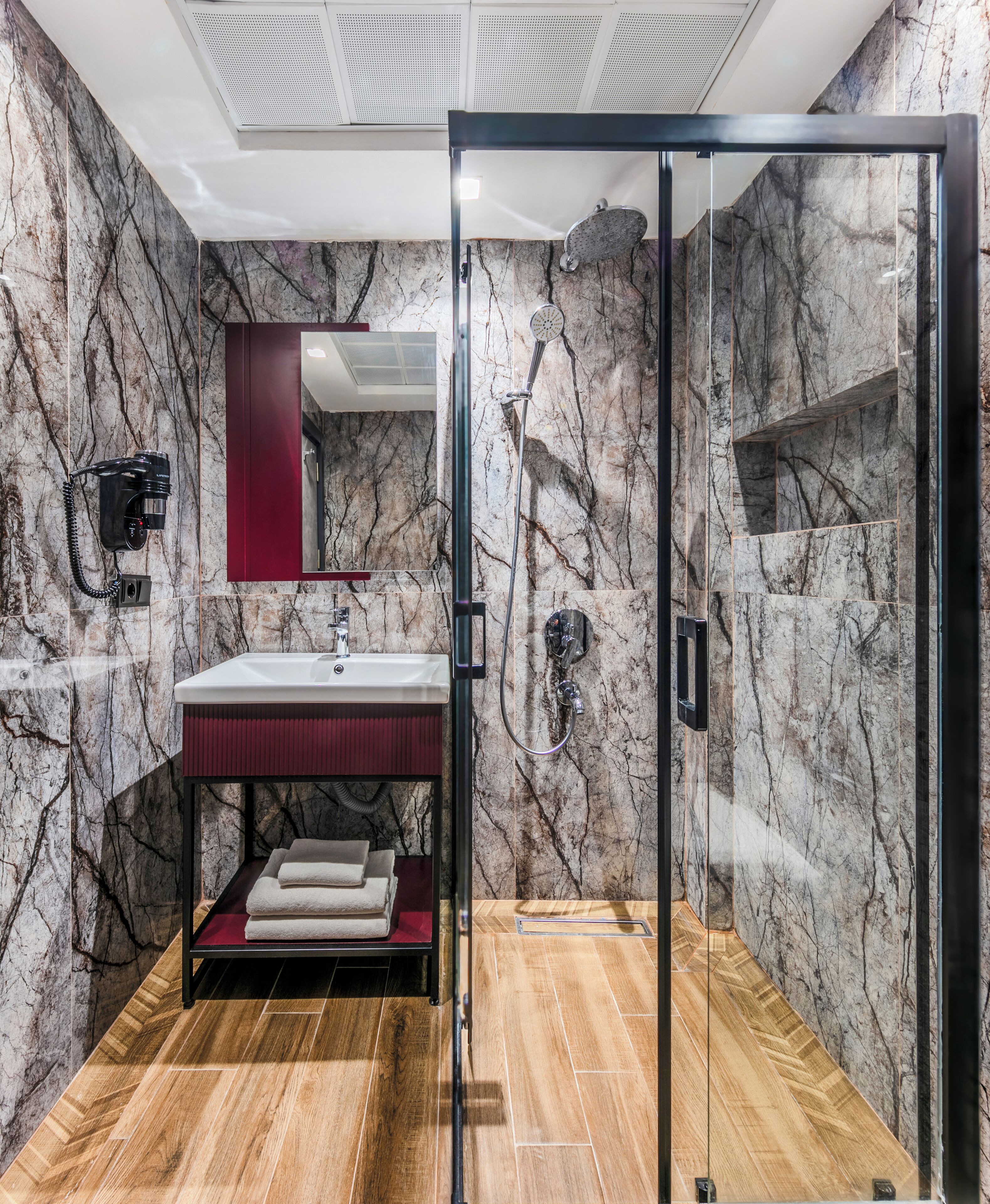Un baño moderno con ducha de vidrio, pared de mármol, suelo de madera y tocador rojo.