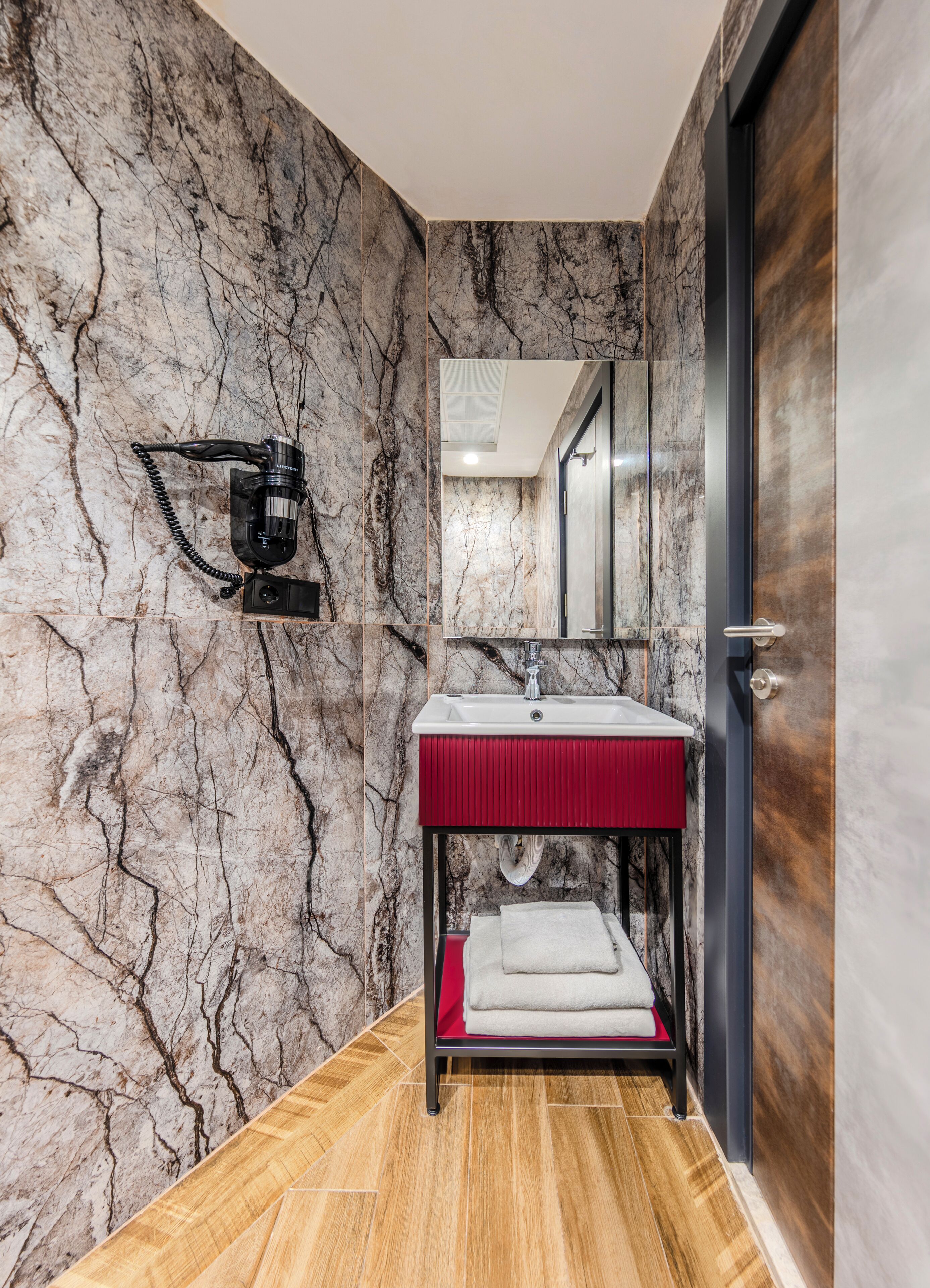 A modern bathroom with a marble wall, a red sink, a mirror, and a wooden floor.