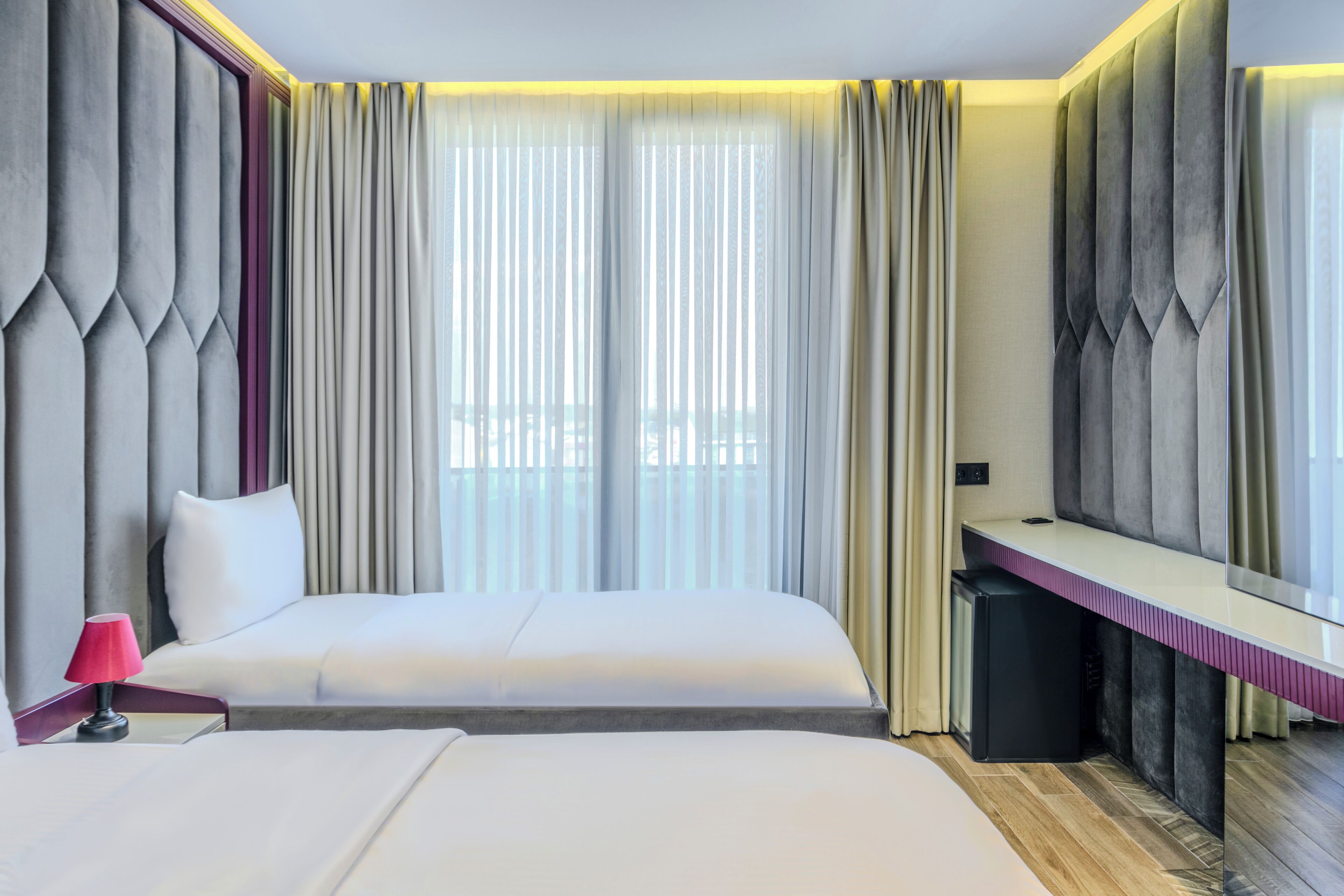 Ein Hotelzimmer mit einem großen Bett, einem Schreibtisch und Blick auf die Stadt durch transparente Vorhänge.