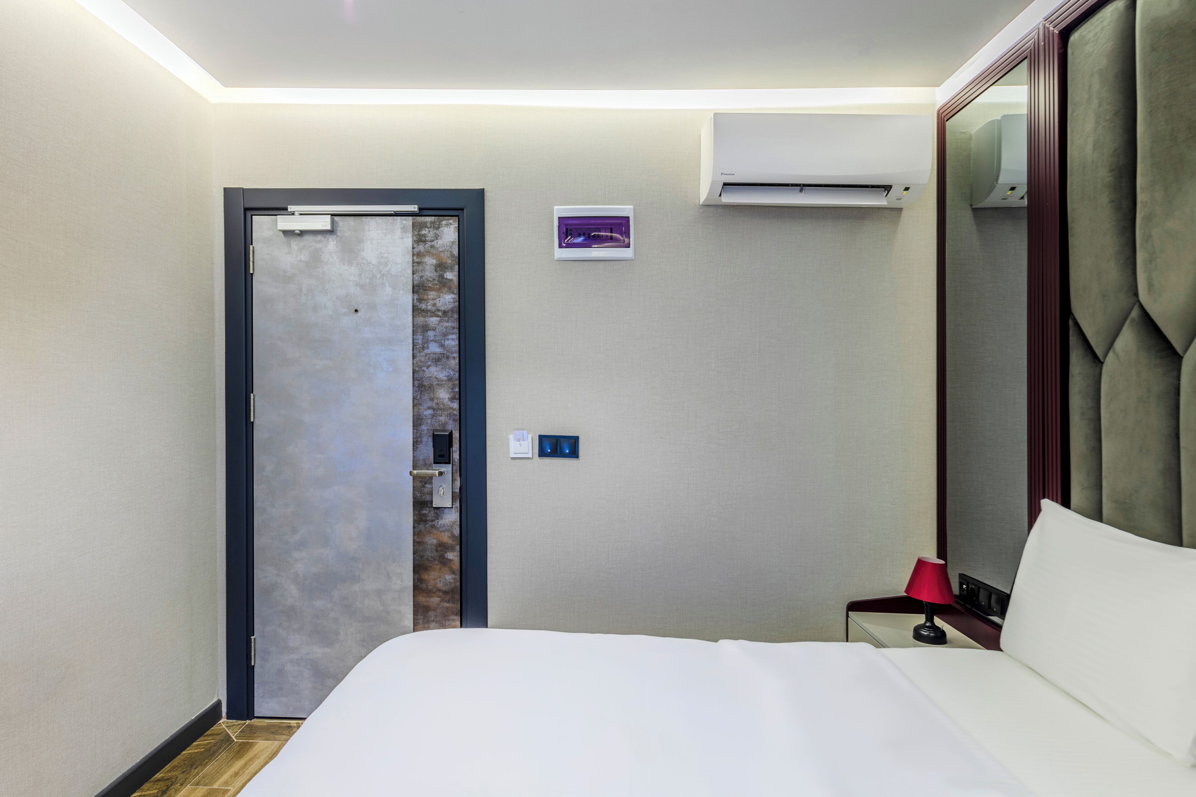 A hotel room with a bed, a nightstand with a lamp, an air conditioning unit, a door with a mirror, and a small wall-mounted control panel.