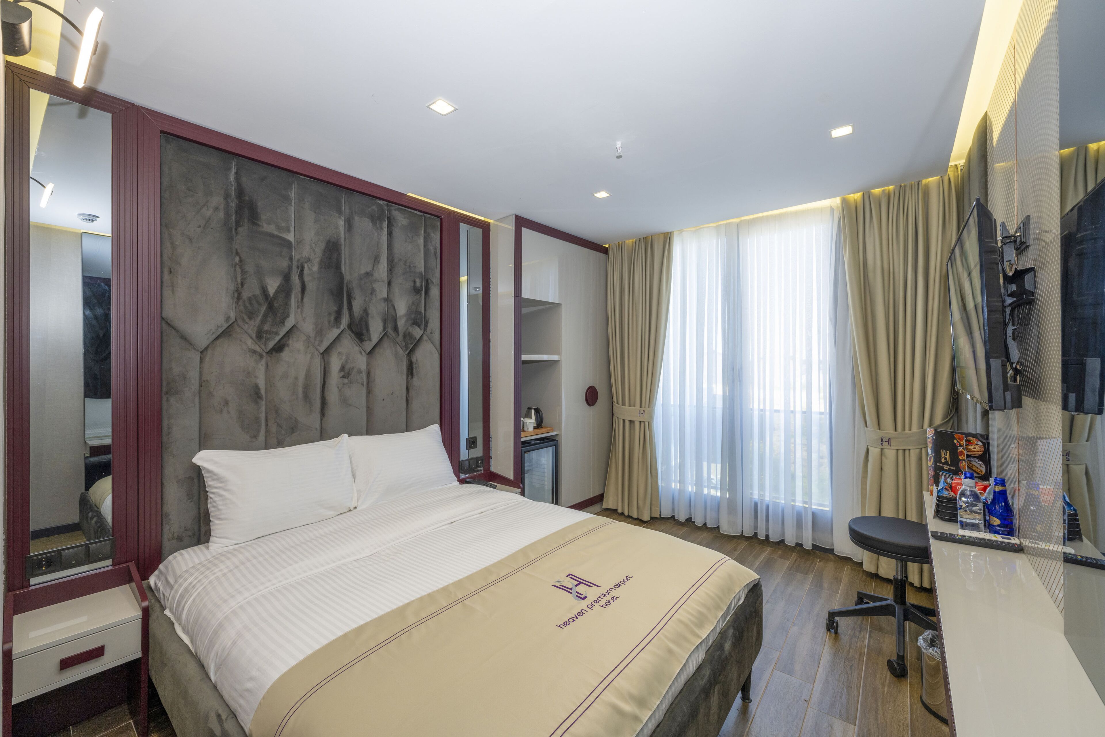 Ein Hotelzimmer mit einem großen Bett, einem Schreibtisch mit Stuhl, einem Spiegel und einem Fenster mit Vorhängen.